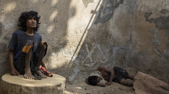 A man sitting on a concrete structure with a chain around his ankle. Another man is lying on the concrete floor.