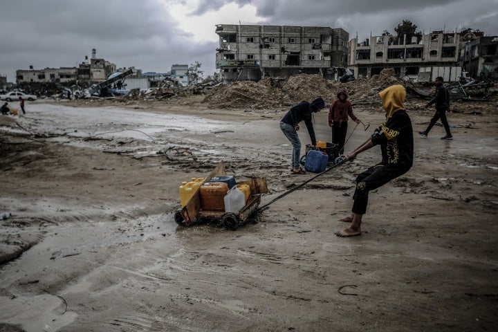 People pull plastic water containers down a muddy street amid destroyed buildings