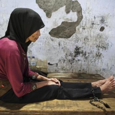 A woman with a scarf sits on a wooden platform to which she is shackled.
